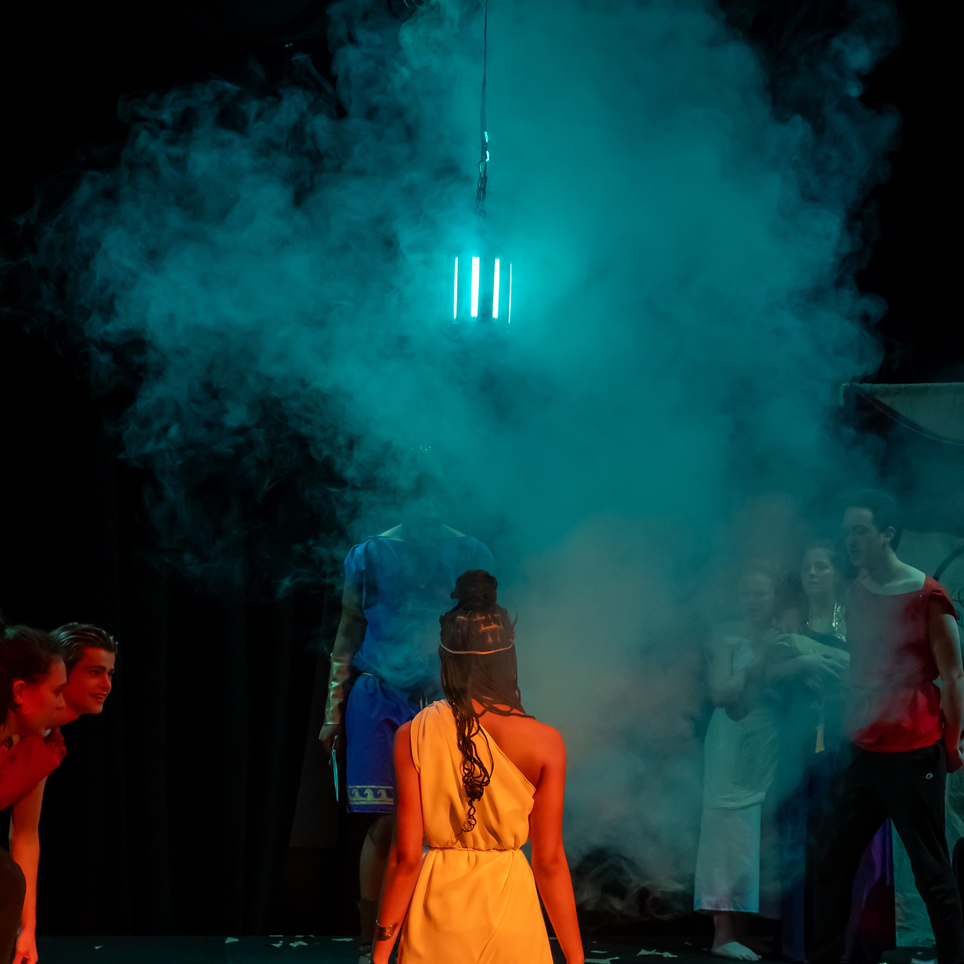A young woman basking in amber light faces away from us, staring at a green lantern in a cloud of fog. Others stare at her, clothed in red.